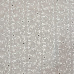 May include: Close-up of a beige fabric with a repeating pattern of white embroidered flowers and vines. The delicate floral design covers the entire surface, creating a textured and detailed appearance. The fabric has a soft, neutral color.