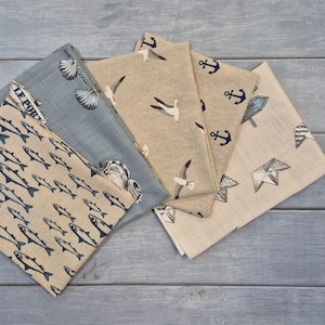 May include: A collection of nautical-themed fabric napkins. The napkins feature various designs, including fish, seashells, seagulls, anchors, and a signpost. The color palette includes shades of blue, beige, and white, creating a coastal aesthetic.
