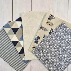 Könnte beinhalten: Eine Auswahl an gefalteten Stoffquadraten mit verschiedenen Mustern. Designs umfassen blau-weiße geometrische Formen, ein neutrales Leinen, ein sich wiederholendes Hausmotiv und ein blau-weißes Wellenmuster. Die Stoffe sind auf einer weißen Holzoberfläche angeordnet.