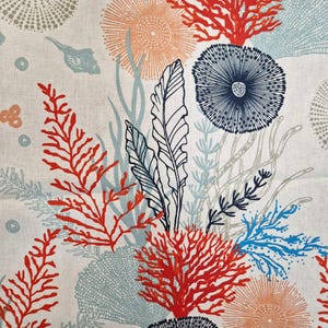 Op de afbeelding: Een stof met een zeethema. Het ontwerp bevat illustraties van koraal, zeewier en andere zeedieren in tinten rood, blauw en oranje. De achtergrond is neutraal van kleur en het kunstwerk is gedetailleerd en complex.