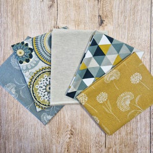 Könnte beinhalten: Eine Sammlung gefalteter Stoffquadrate in verschiedenen Mustern und Farben. Designs umfassen florale, geometrische und abstrakte Drucke in Blau-, Gelb-, Grau- und Weißtönen. Die Stoffe sind auf einer Holzoberfläche angeordnet.