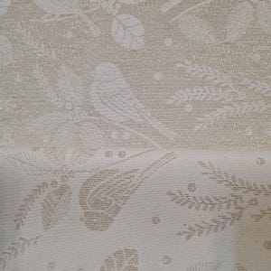 Könnte beinhalten: Weißer Stoff mit einem subtilen Schimmer, der ein sich wiederholendes Muster aus stilisierten Vögeln und Blättern zeigt. Der Stoff hat eine zarte, fast geprägte Textur.