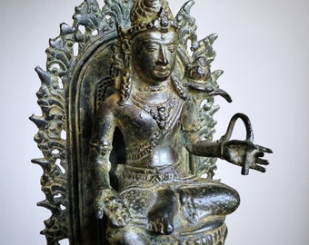 Può includere: Una statua di bronzo di una divinità seduta su un trono con sculture intricate. La divinità indossa una corona ed è adornata con gioielli. La statua è un pezzo tradizionale di arte religiosa.