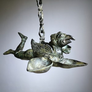 May include: A bronze hanging oil lamp featuring a winged figure with a bird's head. The lamp has three oil reservoirs and a chain for hanging.