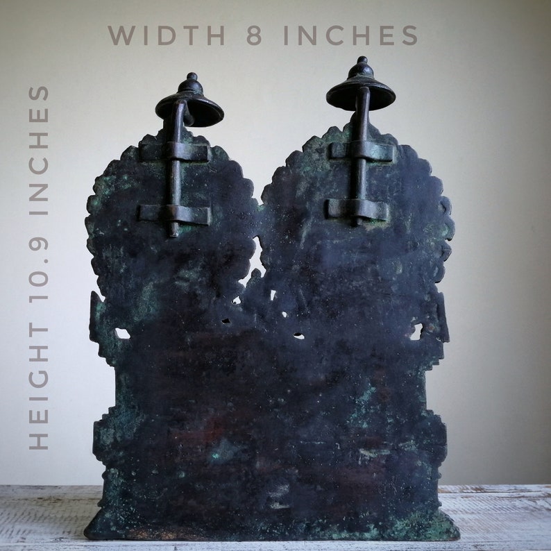 May include: A dark bronze sculpture of two figures, each with a decorative top, standing on a wooden surface. The sculpture is 10.9 inches tall and 8 inches wide.