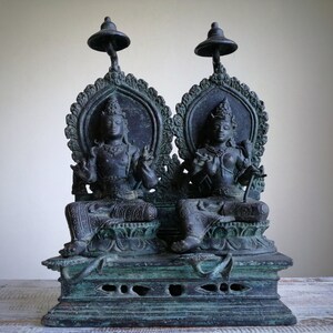 May include: A pair of black bronze statues of deities sitting on a throne with ornate details. The statues are seated on a base with intricate carvings.