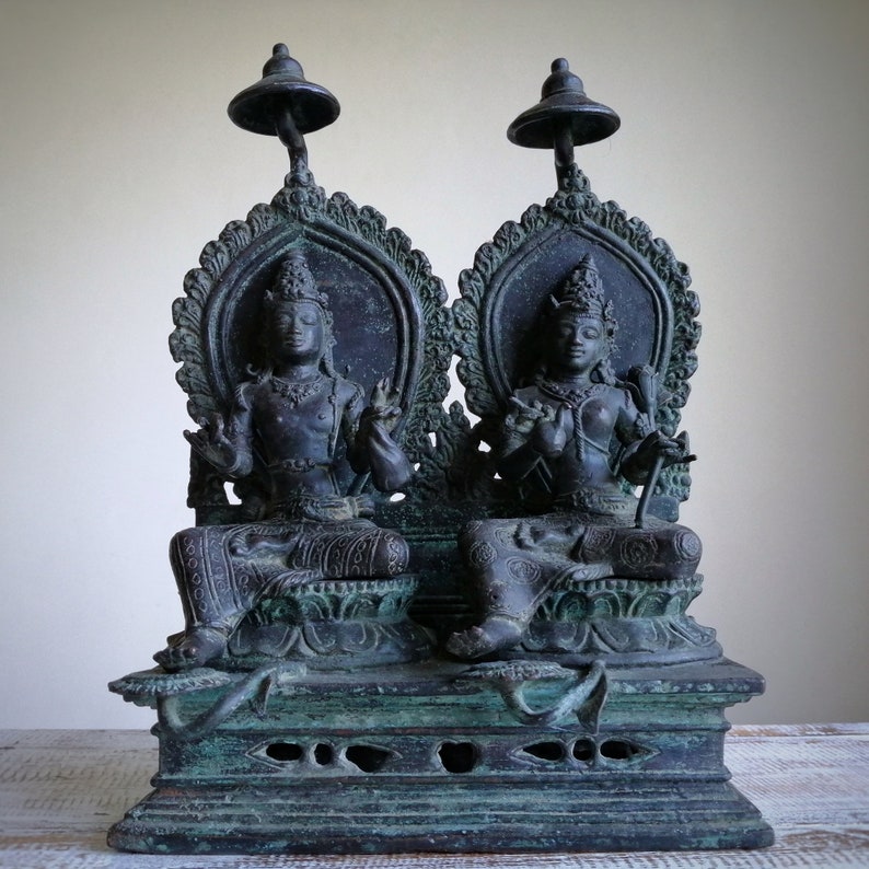 May include: A pair of black bronze statues of deities sitting on a throne with ornate details. The statues are seated on a base with intricate carvings.