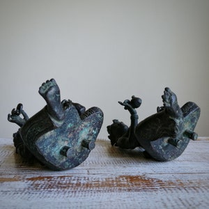 May include: Two antique bronze sculptures of reclining figures. The sculptures are detailed with intricate carvings and have a dark green patina.