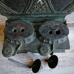 May include: Antique bronze inkwell with two wells and ornate details. The inkwell has two small, round, black inkwells with a decorative design around the rim. The inkwell is sitting on a white surface.