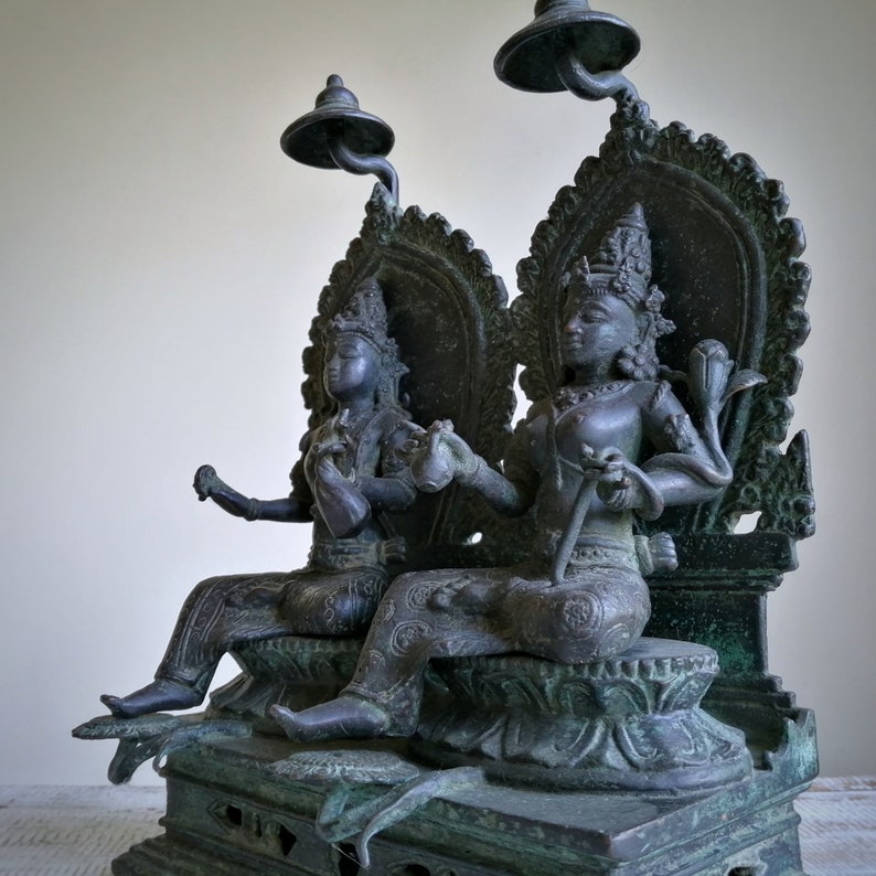 May include: A dark green bronze sculpture of two seated figures, likely deities, with elaborate headdresses and ornate clothing. The figures are seated on a throne-like base with intricate carvings.