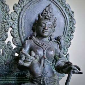 May include: A dark bronze statue of a deity seated on a throne with intricate carvings. The deity is adorned with a crown and jewelry, and holds a small object in one hand.