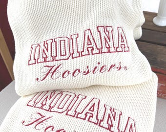 May include: A cream-colored knit sweater featuring a red ribbed crew neckline and red embroidered text that reads INDIANA Hoosiers. The sweater has a thick, chunky knit texture and is folded, showing the same red text on the lower section.