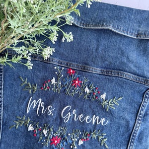 May include: A denim jacket with white embroidery that says "Mrs. Greene" surrounded by a floral design with red, white, and blue flowers.