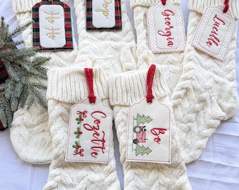 May include: Cream-colored cable knit Christmas stockings with personalized name tags. The tags are rectangular with red or gold ribbon. Some tags have names embroidered in red, others have holiday designs. A small Christmas tree is visible.