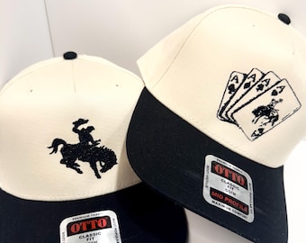 May include: Two baseball caps with black brims and off-white crowns. One cap features a black embroidered cowboy on a bucking bronco, the other has a playing card design. Both have "OTTO" labels.