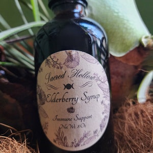 Könnte beinhalten: Eine dunkle Glasflasche mit einem Etikett, auf dem "Sacred Hollow Elderberry Syrup Immune Support Net Wt. 8 Oz" steht. Die Flasche steht auf einer braunen Oberfläche.