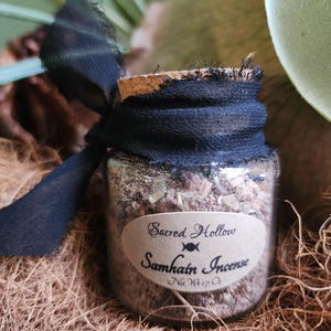 May include: A small glass jar with a black ribbon tied around the top. The jar contains a brown and white incense blend. The label on the jar reads "Sacred Hollow Samhain Incense Net Wt 1/2 oz".
