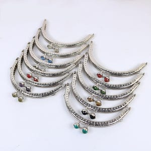 May include: A set of eight silver-toned purse frames with decorative detailing and colorful gemstone accents. Each frame has a half-circle shape and a clasp closure.