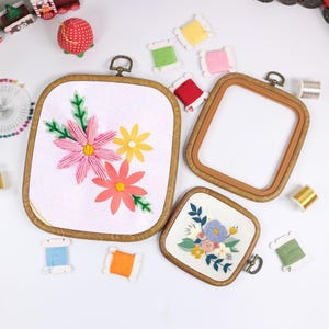 May include: Three wooden embroidery hoops, two with floral embroidery and one empty. The embroidered hoops feature pink, yellow, and coral flowers with green leaves. Spools of thread in various colors are scattered around the hoops.