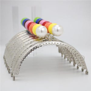 Puede incluir: Un marco de bolso de metal plateado con un borde festoneado decorativo. El marco está decorado con una fila de cuentas de colores y dos acentos de perlas blancas.