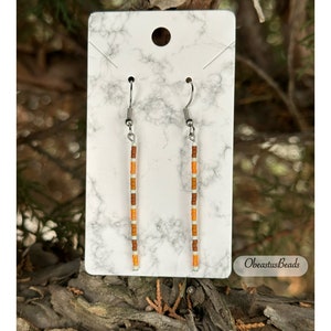 May include: A pair of long, beaded earrings with a brown, orange, and white color scheme. The earrings are hanging from silver hooks on a white marble background.