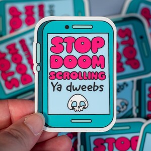 May include: A sticker in the shape of a smartphone with a teal border and a light blue screen. The sticker has the words "STOP DOOM SCROLLING Ya dweebs" in pink and white, along with a skull graphic.