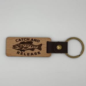 May include: A wooden keychain with a brown leather strap and a brass key ring. The wooden tag is rectangular and features the words "CATCH AND RELEASE" with a fish illustration. The keychain is designed for anglers.