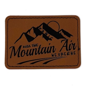 May include: A rectangular brown patch with black text and graphics. The text reads "Kiss the Mountain Air We Breathe" with a mountain and sun graphic. The patch has a stitched border.