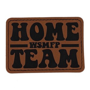 May include: A brown rectangular patch with the words "HOME TEAM" in bold black letters. The text "WSMFP" is centered between the words. The patch has a stitched border and is set against a white background.