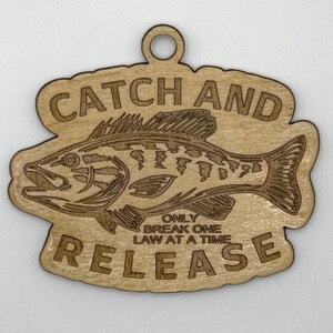 May include: Wooden cutout featuring a detailed illustration of a fish, with the words "CATCH AND RELEASE" and "ONLY BREAK ONE LAW AT A TIME" engraved. The cutout has a small hole at the top for hanging. The fish is a bass.