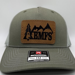 May include: Olive green trucker hat with a brown leather patch featuring a mountain and tree design with the letters "BMFS" in black. The hat has a white mesh back and an adjustable strap.