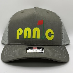 May include: A gray and white trucker hat with a green and yellow "PANIC" logo and a red golf flag. The hat has a black label with the text "RICHARDSON. ORIGINAL 112 TRUCKER ADJUSTABLE."