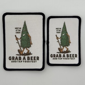 May include: Two white iron-on patches with black borders. The patches feature a cartoon tree character with a smiling face and the text "Meet me at the creek!" and "Grab a beer and tap your feet".