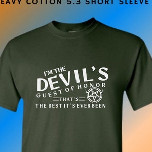 May include: A dark green t-shirt with a white graphic print that says "I'm the Devil's Guest of Honor - That's the best it's ever been". The graphic also includes a pentagram.