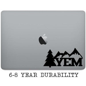 May include: A black and white laptop sticker with a mountain range and two pine trees. The text "YEM" is in the center of the sticker. The sticker is on a silver laptop. The text "6-8 YEAR DURABILITY" is below the sticker.