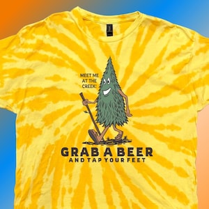 May include: Yellow tie-dye t-shirt with a cartoon tree character holding a walking stick. The text "Meet me at the creek!" is above the tree, and "Grab a beer and tap your feet" is below. The shirt has a crew neck.