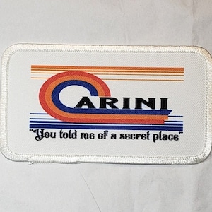 May include: White rectangular patch with a blue, orange, and red striped design. The word "CARINI" is in black letters on the patch. The text "You told me of a secret place" is in black letters below the design.