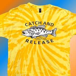 May include: Yellow tie-dye t-shirt with a graphic of a fish and the words "CATCH AND RELEASE" and "ONLY BREAK ONE LAW AT A TIME". The shirt has a crew neck and short sleeves. The design is in black and white.