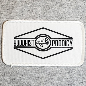 May include: White embroidered patch with black outline of a diamond shape with the words "Buddhist Prodigy" and a megaphone inside.