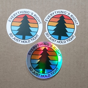 May include: Three round stickers with the text "Everything's Right So Just Hold Tight" and a stylized pine tree graphic. The top two stickers are white with black and blue graphics. The bottom sticker is iridescent with rainbow colors.