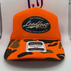 May include: An orange trucker hat with a camouflage brim. The hat features the text "502 Chevelle Leadfoot BMFS Racing" in a navy blue oval. The brim has a camouflage pattern with brown and green shapes. The hat also has a "OTTO" label.