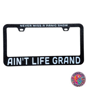 May include: A black glittery license plate frame featuring the white text "NEVER MISS A PANIC SHOW" along the top edge and the white text "AIN'T LIFE GRAND" along the bottom edge. The frame has two mounting holes at the top for vehicle attachment.