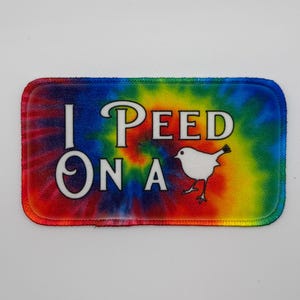 May include: Rectangular patch with a tie-dye background in shades of red, blue, green, and yellow. The patch features the text "I PEED ON A" in white, with a white bird illustration.