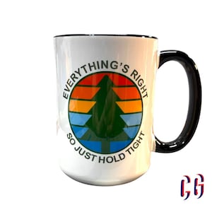 May include: A white ceramic mug with a black handle and rim. The front features a circular graphic with a green pine tree silhouette against a sunset-colored background of orange, yellow, and blue stripes. The text reads Everything's Right So Just Hold Tight.