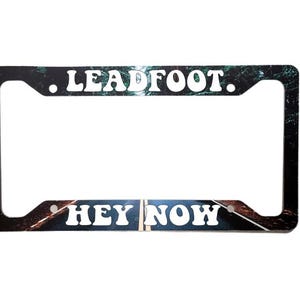 May include: A black license plate frame with a road scene and the text ".LEADFOOT." at the top and "HEY NOW" at the bottom.