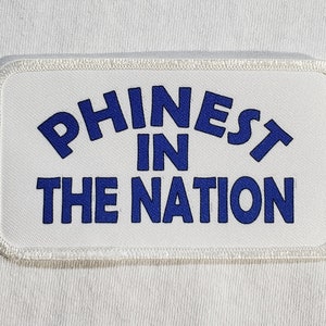 May include: White rectangular patch with blue embroidered text that reads "Phinest in the Nation".