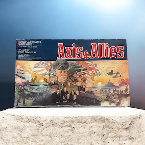 May include: The Axis & Allies board game box features a dark blue top section with the title in red and white text. The illustration depicts military figures, tanks, ships, and aircraft against a backdrop of world maps and national flags.