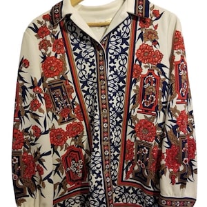 Puede incluir: Camisa abotonada de manga larga con fondo crema y estampado floral en rojo, azul marino y marrón. La camisa tiene cuello y puños blancos y está colgada de una percha de madera.