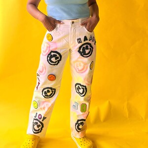May include: White denim jeans with a tie-dye effect and various patches featuring smiley faces and the words "HAPPY" and "JOY".