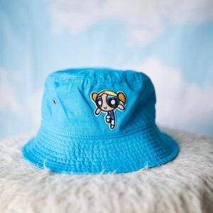 May include: A blue bucket hat with a cartoon character embroidered on the front. The character is a girl with blonde hair and a blue dress. The hat is on a white fluffy surface.